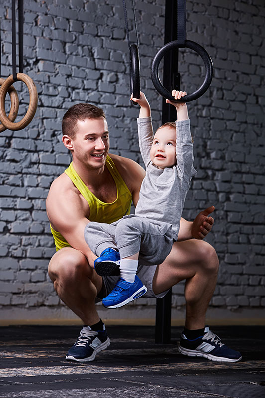 Happy father and little son exercising with gimnastic rings and smiling against brick wall in the gym. Fathet and son in the sportwear and sportive shoes. Happy family. Rearing child in the healthy lifestyle.