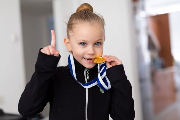 Portrait of a little girl-gymnast in a tracksuit with medals on the neck, biting the medal and showing thumbs up